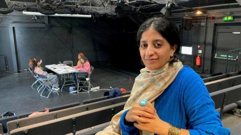 A woman wearing a blue jumper and yellow scarf with four people sitting around a table in the background