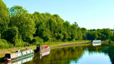 view of a canal with three canal boads with lots of green trees on the banks.  Blue skies and sunshine above