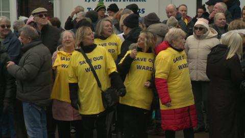 Five women in yellow shirts with slogan saying "Save our ward" stand in middle of crowd of protesters outside the hospice