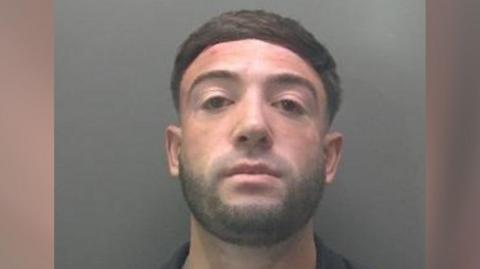 Custody image of Dominic Bevilacqua - he is looking into the camera with his chin raised. he has black hair and a short stubble beard