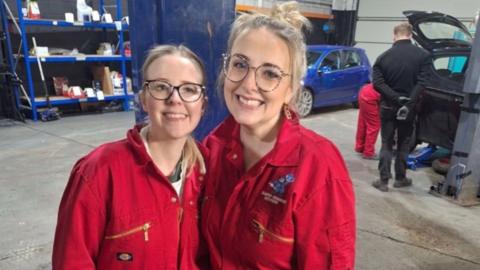 Two women stand smiling in a car garage. They are both wearing red overalls. There is a blue and black car behind them, with two people looking at one of the cars.
