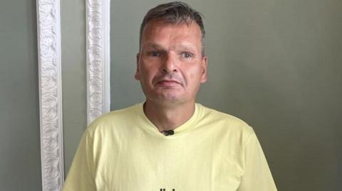 A man, William Fleming, in a yellow t-shirt stood in front of a green wall with white moulding. He has a serious expression.