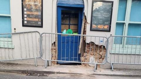 The entrance to the pub fenced off, with the wall and door damaged.