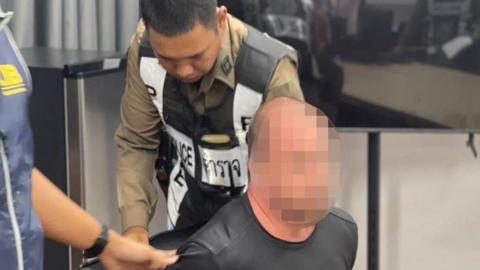 A blurred face of a man wearing a black top with this hands behind his back as he is being arrested. A Thai police officer is stood behind him 