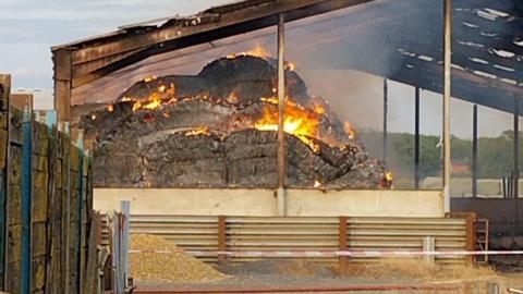 A barn fire, showing flames, on hay, with its roof burnt. There is red tape across the front, a fence to the left and barriers in front. 