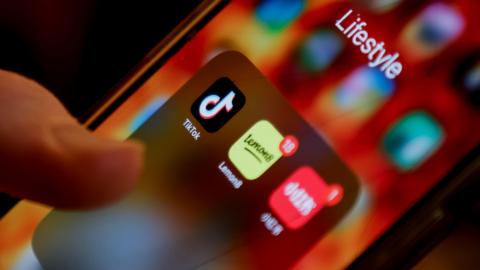 A close-up image of a mobile phone. A thumb is seen touching a screen displaying the TikTok logo.