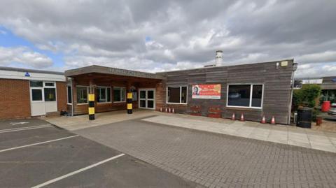 A red brick building with horizontal wooden panels on half of the building. Writing above the doorway of the building reads 'Humberston Park School.' There is pavement and car parking spaces in front. There are light grey clouds in the sky.