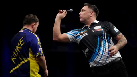 Gerwyn Price, pictured throwing a dart at a dartboard out of shot. He is wearing a black shirt with blue and white shards across it and a white collar. Luke Littler has his back to the camera, looking straight ahead. The background is black. 