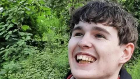 Roan Bloore outdoors in a lush, green environment. The background is filled with dense foliage, including various shades of green leaves and plants. He is wearing a dark-coloured garment with a lighter stripe and a visible red inner lining near the collar.