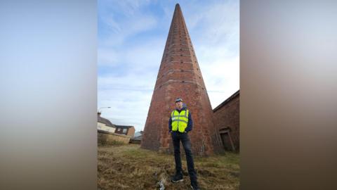 Rafferty Specialist Access Services' contracts director Carl Woodroffe standing at the bottom of Dixons Chimney in Carlisle. He is wearing a hard hat and yellow hi-vis vest, over a blue jacket and black jeans. He is looking down at the camera. Dixons Chimney is a red-brick structure with a hexagon shape. There are metal belts around the structure.