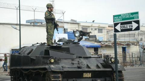Two Ecuadorean soldiers stand guard atop a tank outside Machala prison, in Machala, Ecuador on January 15, 2024. One is carrying a weapon, and in the background the wall of the prison can be seen with fencing and barbed wire on top.
