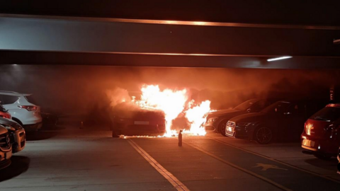 A car is on fire inside a multi-storey car park. It is not parked in a bay. There is an abandoned fire extinguisher on the floor. 