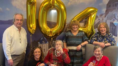 A family sitting in a row in front of birthday balloons in the shape of the numbers 104. In the centre is a woman with a breathing apparatus - her family smiles at the camera.
