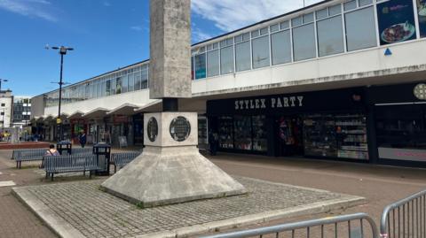 A row of shops in Harlow with a memorial statue in the foreground and metal benches in the background.  The block of shops are one storey and the main colours are beige and grey.