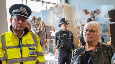 A man in a high visibility police jacket and police cap and a woman in a black leather jacket talk to two men. 