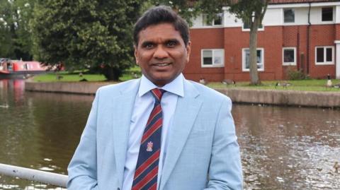 Dr Chandra Kanneganti wears a pale blue suit with a stripy blue and red tie. He is smiling, and posing next to the canal in Stoke-on-Trent, with a narrow boat in the background, along with waterside properties. 