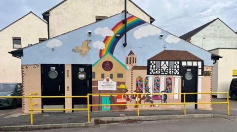 The front of a toilet block has been painted with images of the old black and white pillared town hall, St Bartholomew's church with stained glass windows and a Great Bustard flying in the sky. Cartoon animals can be seen in the foreground. And a large rainbow painted above.