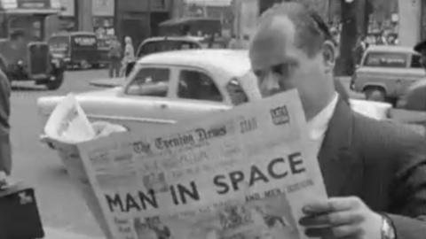 Man wearing suit reads the Evening Times on the side of the street. Headline on newspaper reads: MAN IN SPACE.