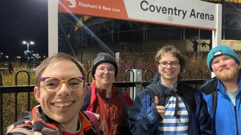Four young men at Coventry Arena Railway Station. Stood in front of the station sign on the platform is a man with yellow and purple glasses, a man with red coat, glasses and black hat, a man wearing glasses with a blue coat with blue and white striped top, and a man with a blue jacket on, ginger beard and turquoise beanie hat 