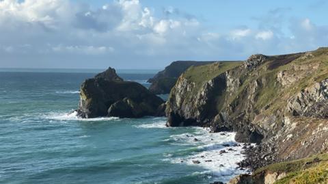 A shot of the coast near Lizard Point in Cornwall. On the left there is blue sea with a large rock in the distance. On the right is a cliff which is green and grey. There are waves crashing up against it. 