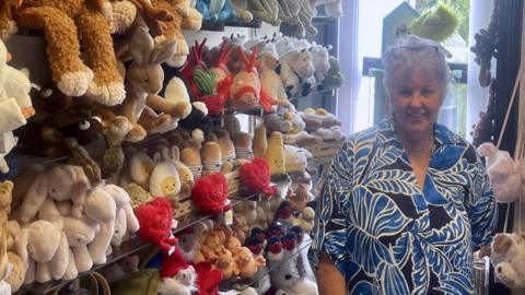 A woman with short white hair, a blue and white patterned shirt, and glasses on the top of her head stands, smiling, next to a large display of Jellycat plush toys including rabbits and fish