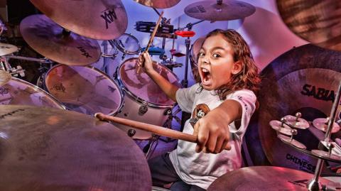 A child with long hair and a white t-shirt drumming