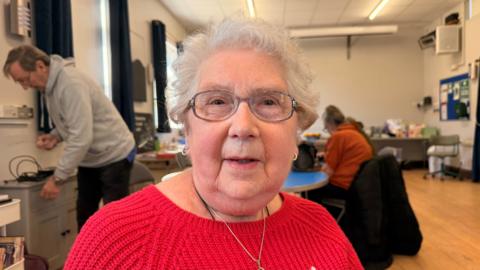 A lady in a red jumper with glasses and white hair. She's in a village hall at a coffee morning. She's elderly and wearing nice earrings and a necklace. 
