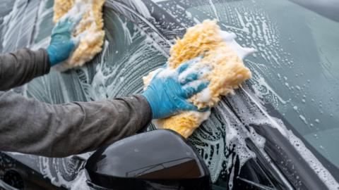 Two blue-gloved hands holding yellow sponges washing the window of a black car.