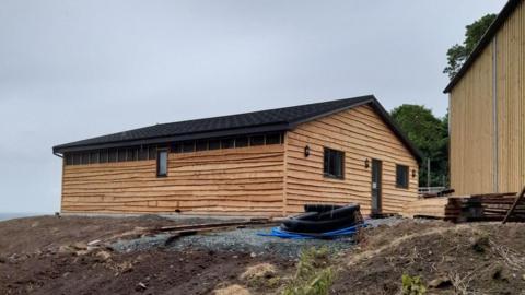 A wooden lodge with a pitched roof is built on land with bits of pipe dumped next to the structure.