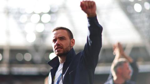 Liam Payne holding his fist in the air standing in a football stadium. He is wearing a blue jacket, a white t shirt and a lanyard sits around his neck.