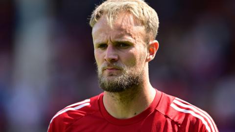Lewis Ward is wearing a red goalkeepers top with white stripes on the shoulders. He has a blonde and brown beard.