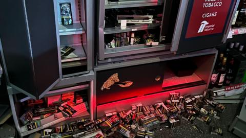 Dozens of cigarette cartons are spilled on the floor under a vending machine that has been broken into by shoplifters