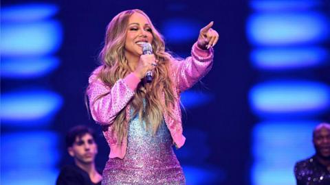 A woman in a pink jacket. She is performing on stage. There are male performers in the background. 