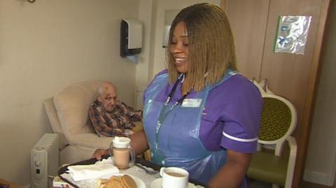 Charity Lawrence, a health care assistant in a care home, wears a purple uniform and holds a tray of tea and biscuits. She has shoulder-length braided hair and is smiling. Behind her is an elderly man in a chair.