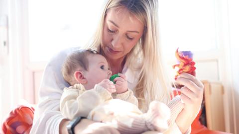 Hayley O'Sullivan holding one of her young daughters. Mrs O'Sullivan has long blonde hair and is wearing a white shirt. She is holding a red toy dinosaur in her left hand. Her baby is wearing a cream outfit and has a green dummy in her mouth.