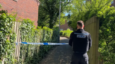 The back of a police officer in front of some blue police tape with a path behind the tape. The path goes between two fences with trees overhead.