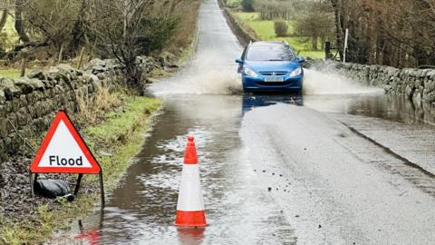 A blue car driving through a small flood in a country road. There is a hazard sign warning of the flood and a cone in some standing water. 