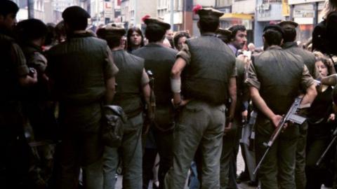 It is 1972. Soldiers stand with their backs to the viewer. One of the soldiers on the right of frame is carrying a large calibre gun. Faces of people in the crowd beyond the soldiers can be seen. In the background, a city centre street.