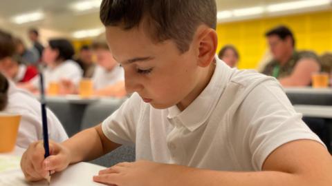 A 10-year-old boy in a white T-shirt is engrossed in writing with a pencil and paper. You can see other children in the background.