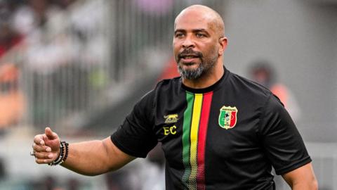 Eric Chelle wears a predominantly black Mali training top, with green, yellow and red stripes on the front and his initials on it as he stands on the touchline during a football match