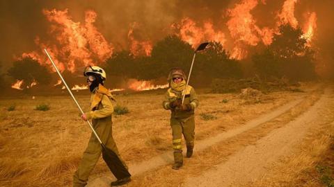 Two female firefighters tackle a raging inferno in a Spanish woodland scene. The grass around them is dry and parched, and the trees in the background are completely aflame.