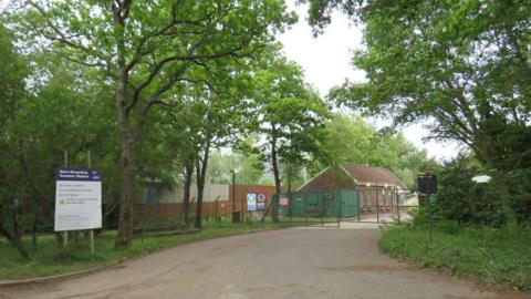 Stock photo of the entrance to the recycling station. There is a red-brick reception building with a pitched roof behind metal green gates.