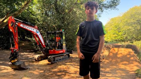 Flynn stands on the bike park in front of a bulldozer. The area is surrounded by dense green woodland, with large trees arching overhead and casting dappled shadows across the ground.