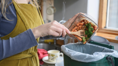 A woman wearing a mustard coloured apron, scraping food waste into a green food caddy