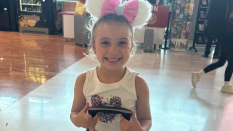 A close-up of Savannah. She is smiling while holding the handle of her suitcase. She is wearing a pink and white bow and a white vest with a heart pattern on it.