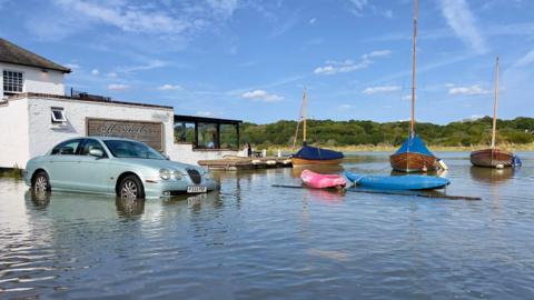 A car parked outside a pub on a flooded road with boats in the distance