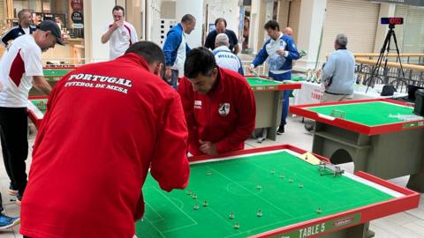 Several men in different coloured tracksuits standing round waist-height tables which have green felt football pitches on them. The pitches have small goals and players.