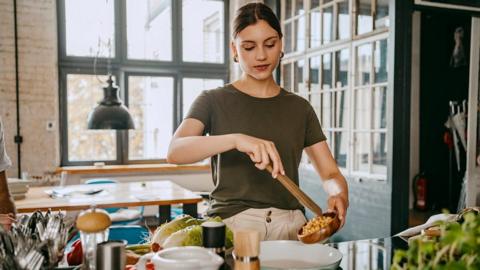 A woman in a kitchen wearing a khaki t-shirt cooking chickpeas on the stove