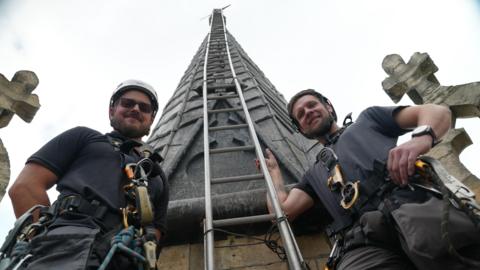 Two men in black tops and shorts are wearing white helmets and harnesses. The photo is taken from the floor looking up. They are holding on to a ladder which ascends up a grey pinnacle with a weather vane at the top.