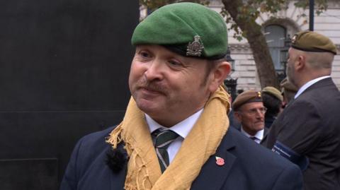 A man with a brown moustache is wearing a green beret, a sand coloured Shemagh Scarf around his neck, and a navy suit with a black tie and poppy badge. He is looking off to the side as he is being interviewed for BBC television news. 